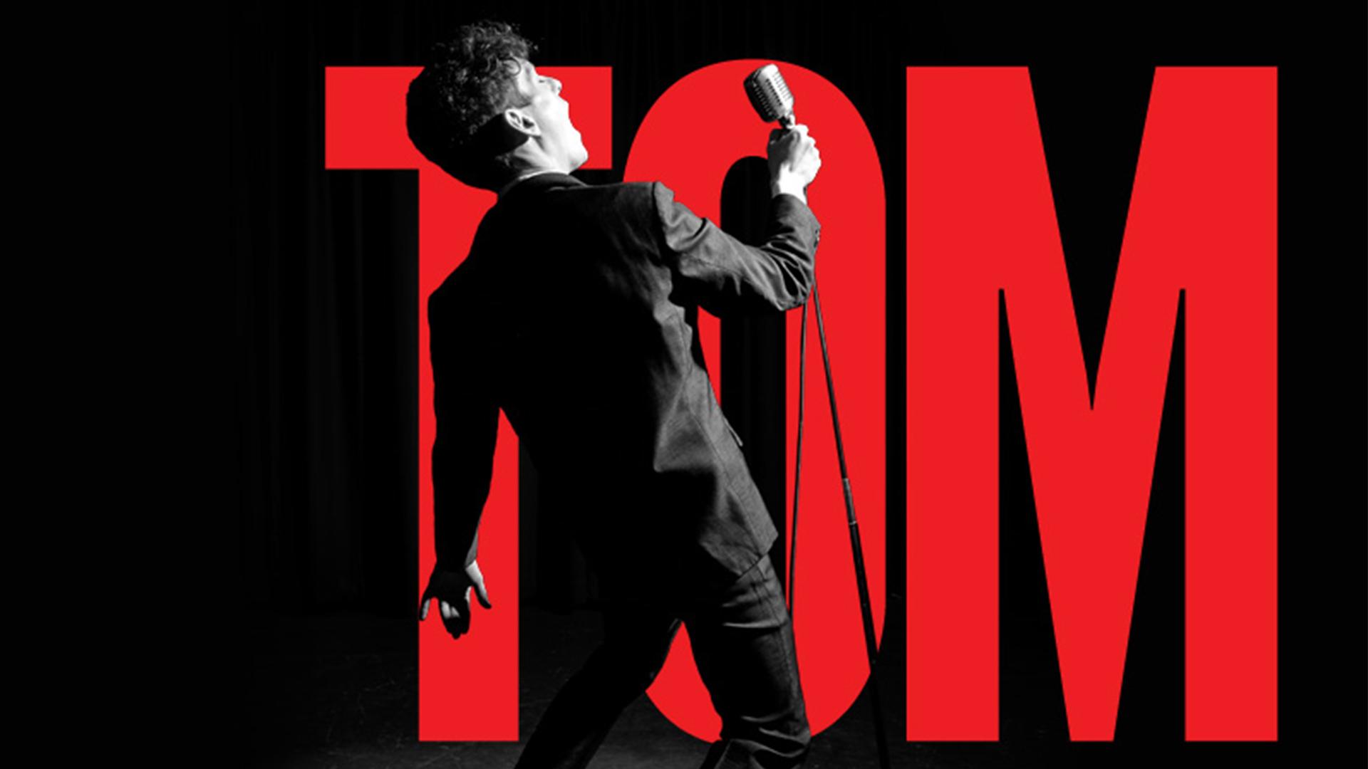 A man holding a microphone on a stand with his back to us with the word TOM in large letters
