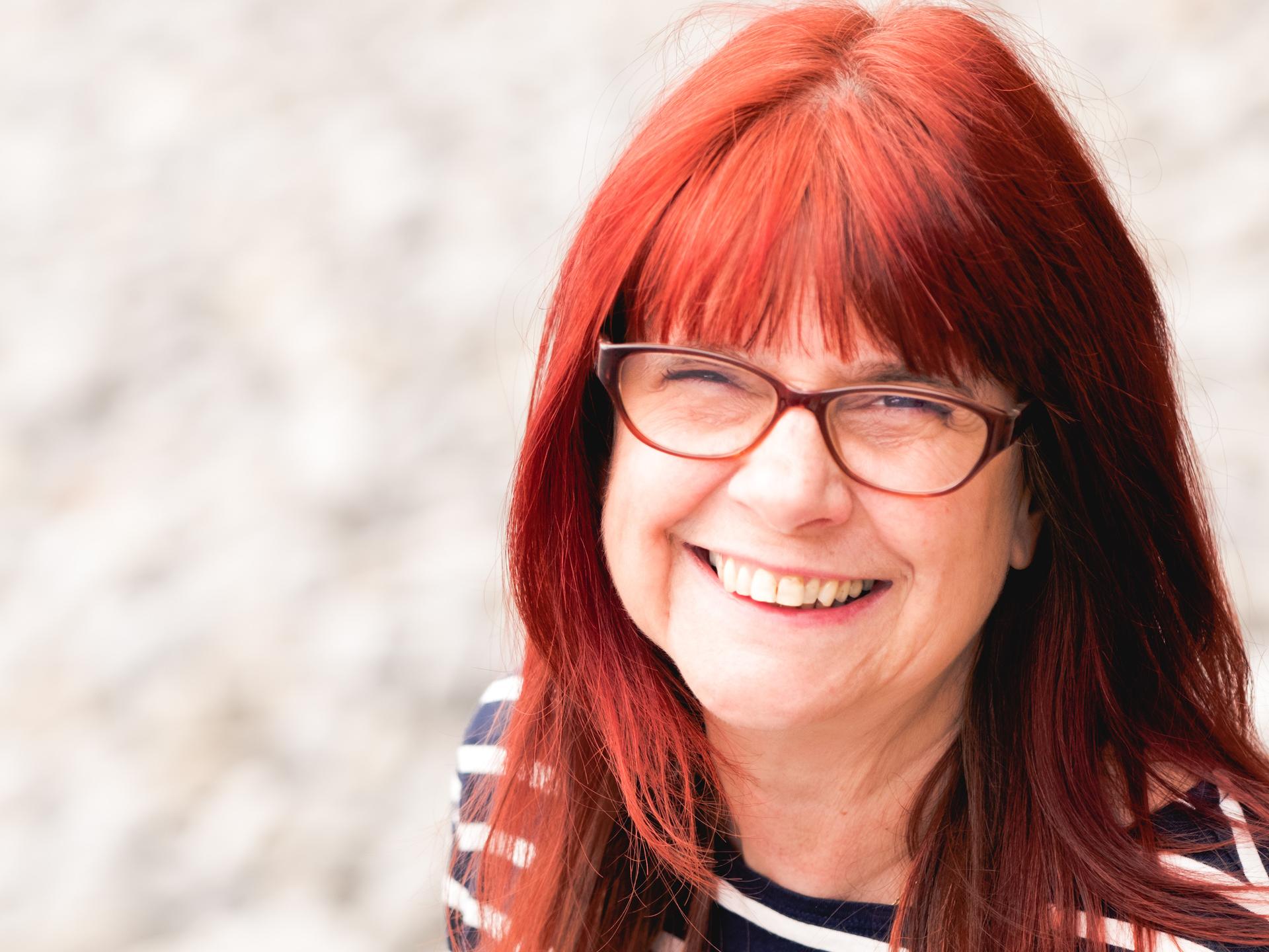 A woman with red hair and glasses wearing a black and white striped top smiling 