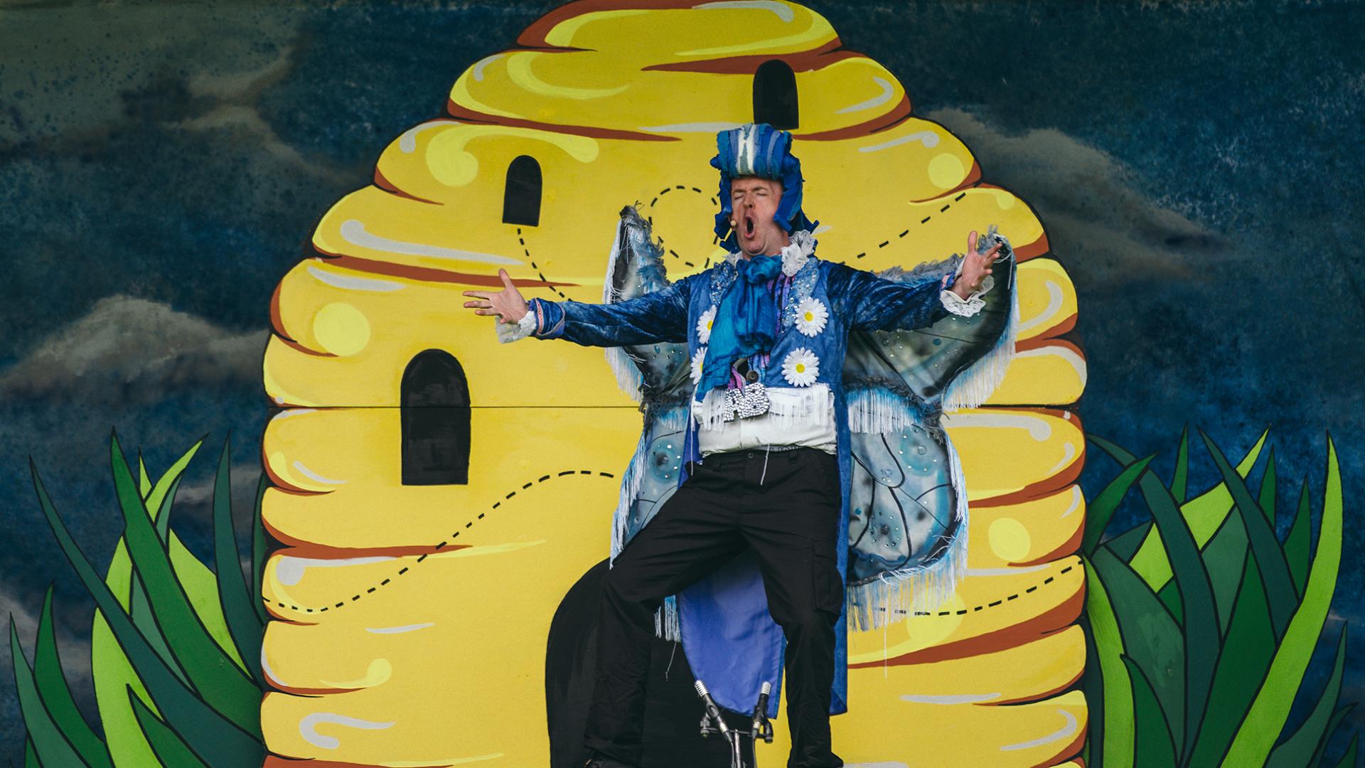 A man dressed as a blue butterfly standing in front of a stage set of a beehive.