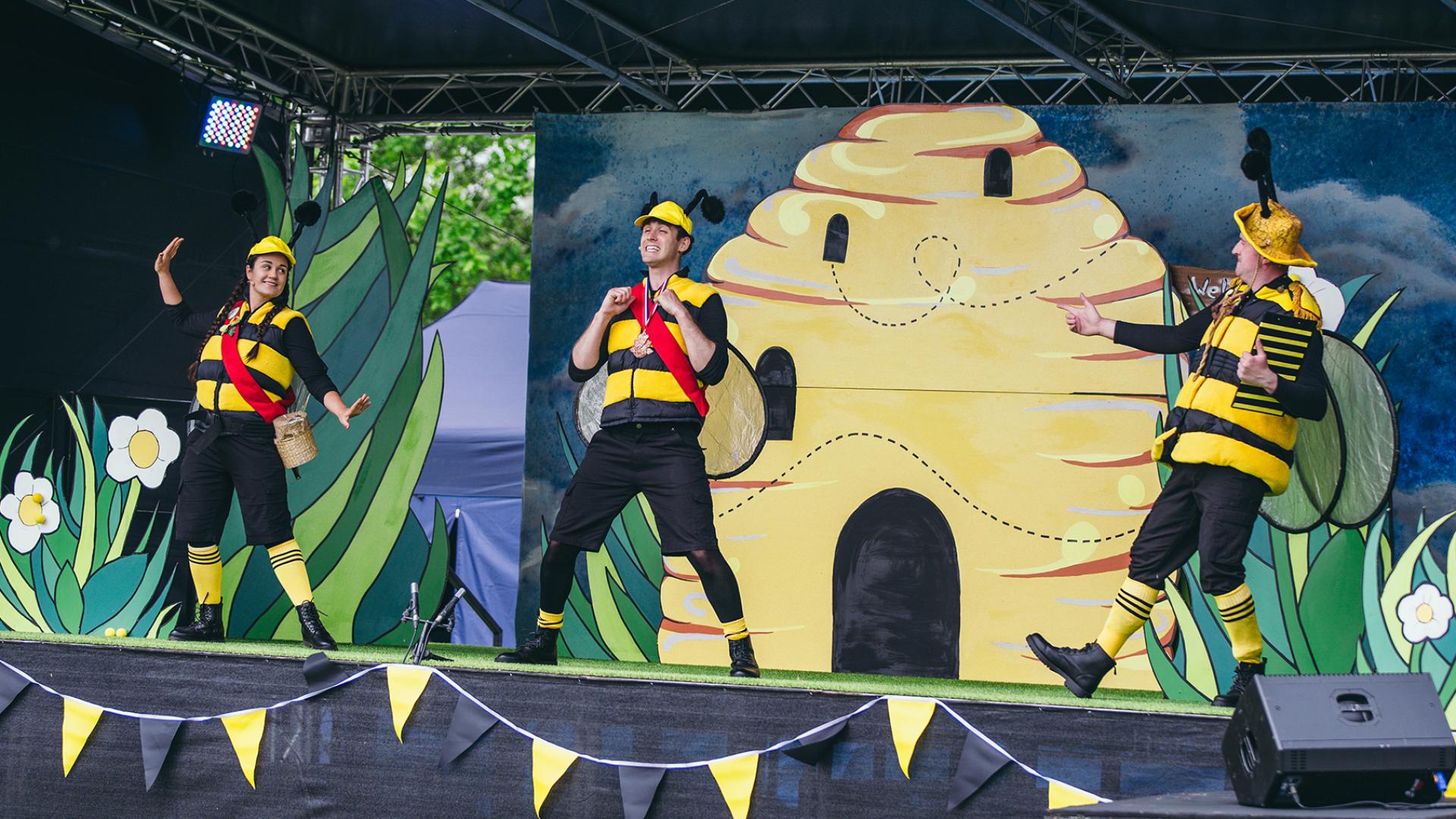 Three people dressed as bees dancing in front of a stage set of a bee hive.