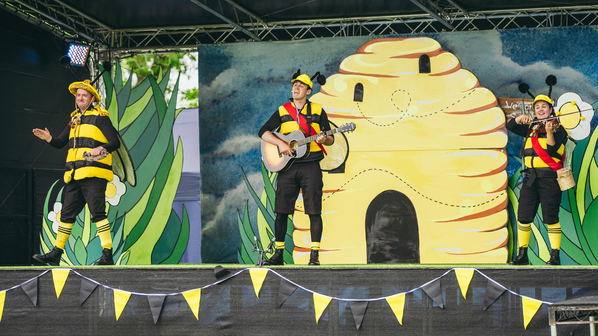 Three people dressed as bees, one playing the guitar, one the violin. All standing in front of a stage set of a bee hive.