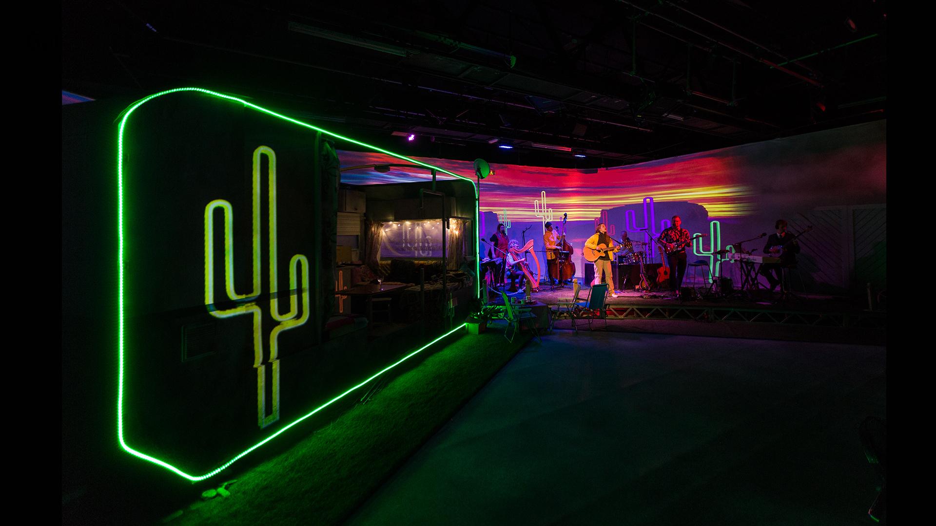 The outline of a caravan and a cactus in green neon light in the foreground with a band in front of a colourful sunset 