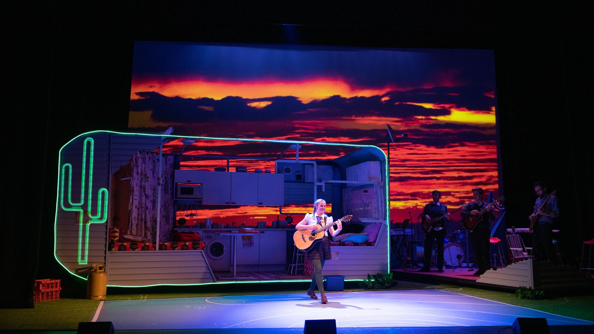 Set of an open sided caravan with a neon cactus, colourful sunset and a school girl playing the guitar