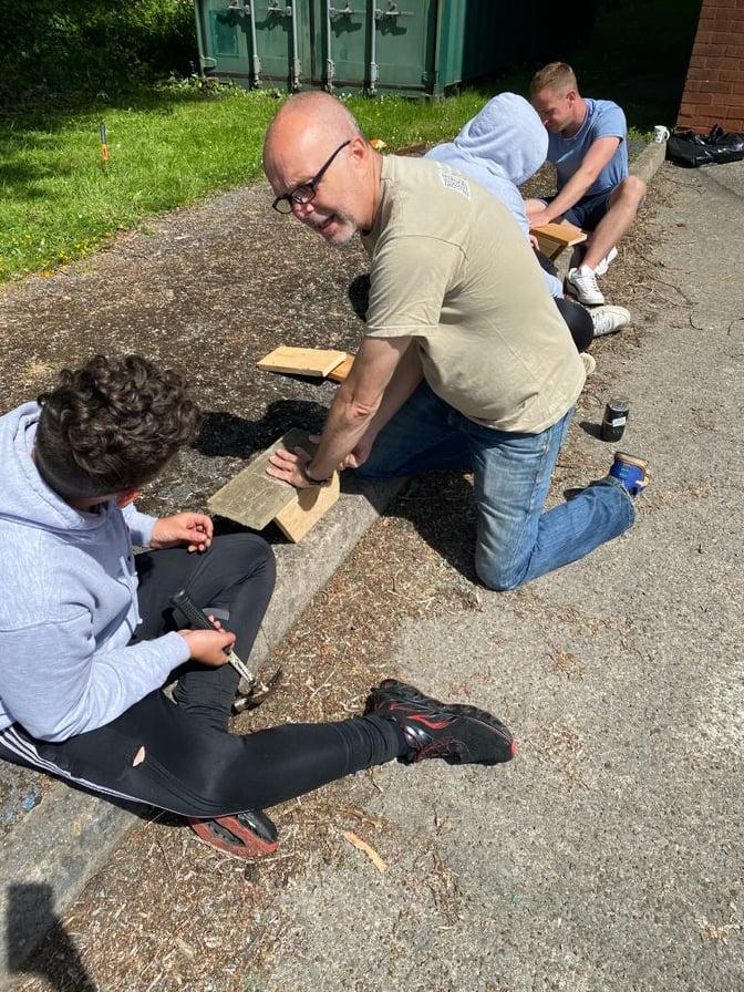 A man with glasses, a green top and blue jeans helps a boy with a grey hoodie and black trousers to create a bird box out of wood