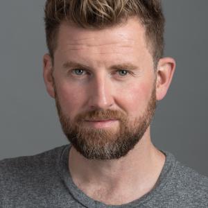 Man with brown hair and a beard wearing a grey top