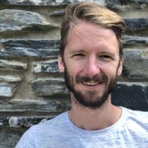 Man with blonde hair and facial hair smiling in front of a wall wearing a grey top